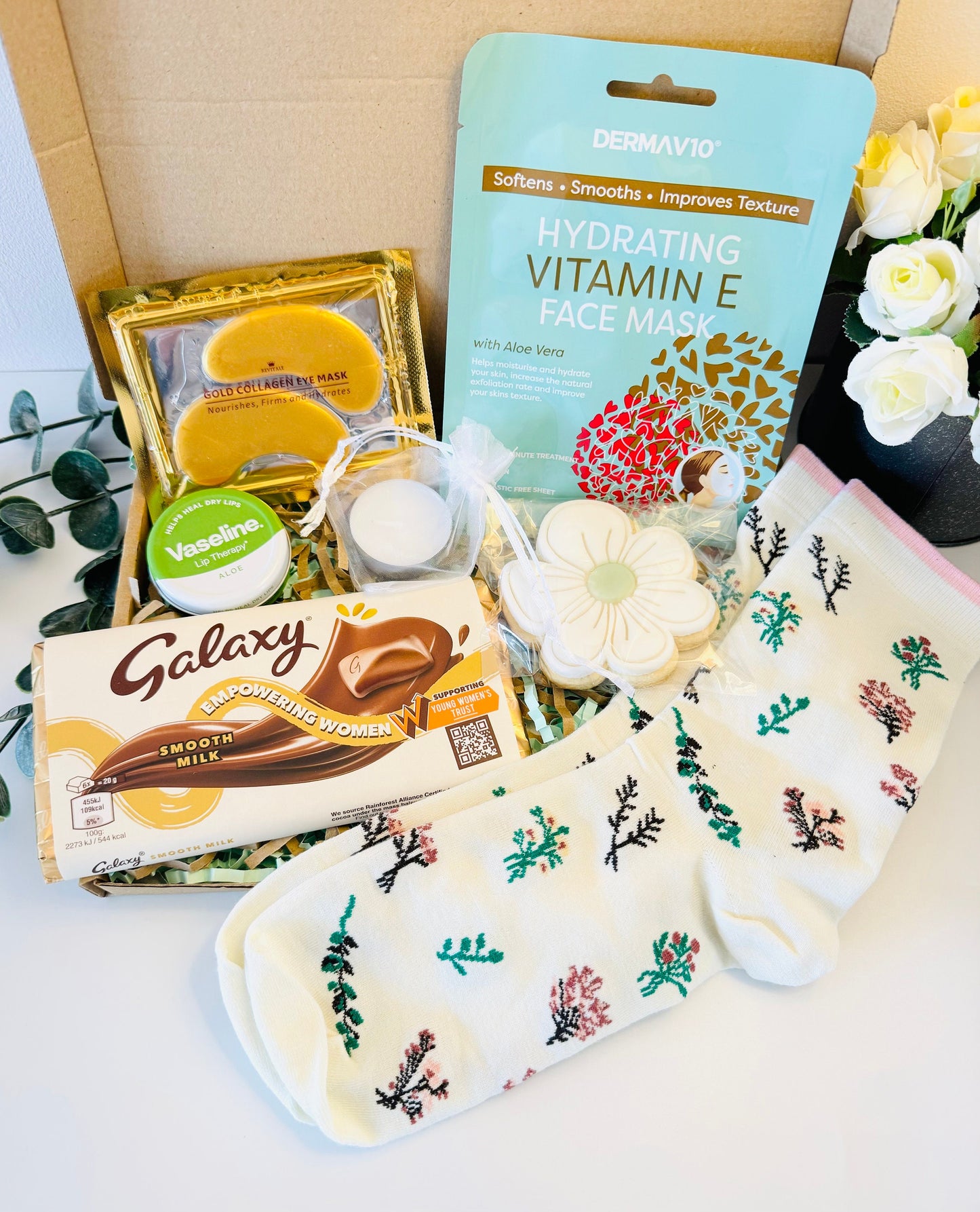 : A gift box filled with various self-care items including a white face mask with the text 'DERMAV10 Hydrating Vitamin E Face Mask with Aloe Vera', a gold collagen under eye mask which Nourishes, Firms, Hydrates', a tea light candle, Vaseline Lip Therapy, a Galaxy chocolate bar, a flower iced biscuit and a pair of white socks with a floral pattern. Comes with a choice of gift card options and a personalised message delivered straight to the recipient’s door