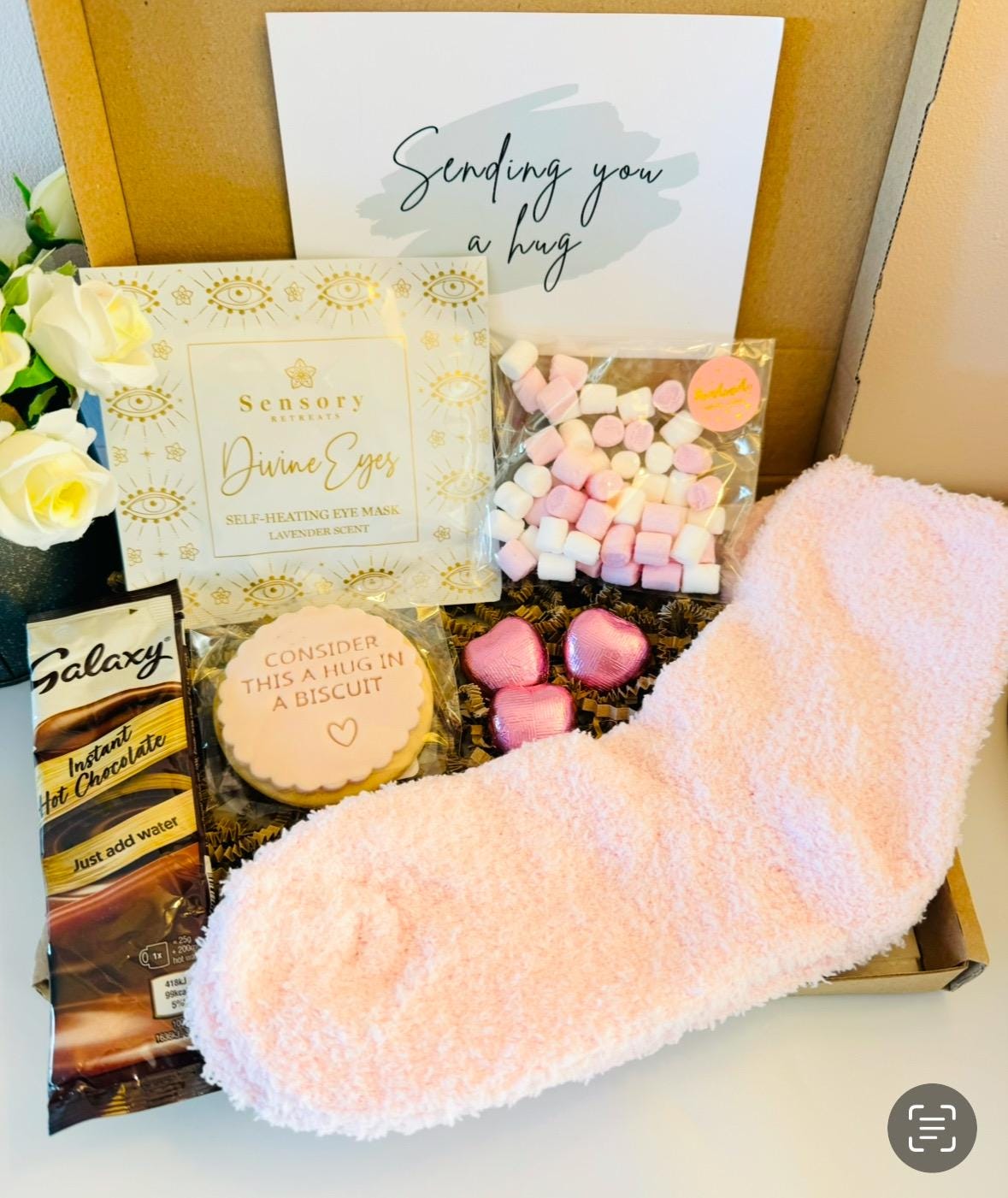 Letterbox Gift filled with pink fluffy socks, 3 mini heart chocolates, galaxy hot chocolate sachet, a self heating eye mask perfect for tired eyes and busy minds, a sending you a hug iced biscuit and a small bag of mini marshmallows. With the option to select from a gift card and add your own personalised message. Delivered straight to the recipient