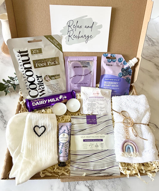May include: A cardboard gift box containing a coconut water foot pack, purple eye masks, a blueberry clay mask, a Cadbury Dairy Milk chocolate bar, two white tea light candles, a lavender bath bomb, white socks with a black heart, scented hand cream, a self-heating eye mask, a white cotton flannel, and a rainbow keyring. With a choose of gift cards, with your own personalised messsge ready to send direct to the recipient