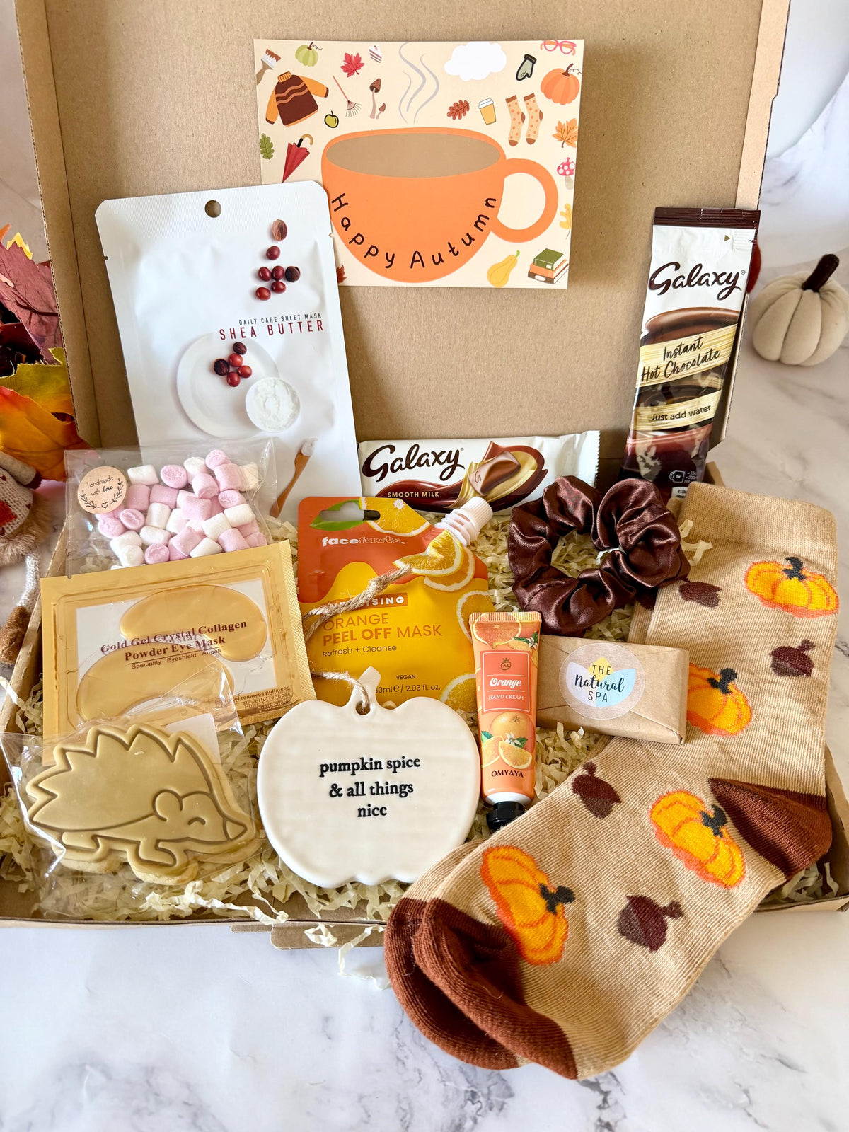 May include: A gift box containing a shea butter sheet mask, a packet of marshmallows, gold collagen eye masks, a hedgehog-shaped biscuit, an orange peel-off mask, hand cream, a satin hair scrunchie, a mini travel soap, a Galaxy hot chocolate sachet, a chocolate bar, and pumpkin & acorn themed socks. Includes a personalised message ready to send direct to the recipient